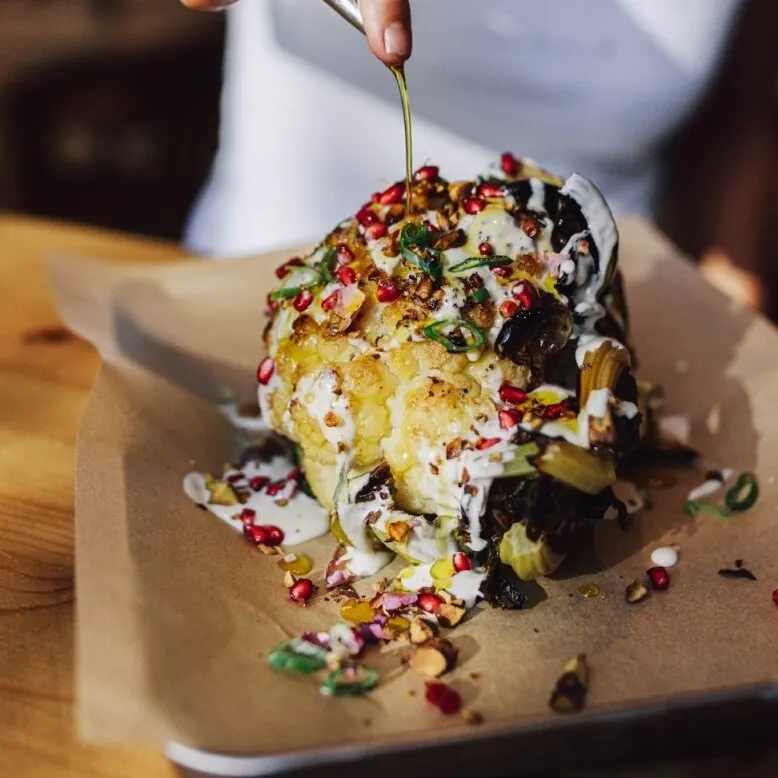 Persona vierte aceite sobre coliflor asada con granada, hierbas y salsa cremosa, servida sobre papel marrón en bandeja.