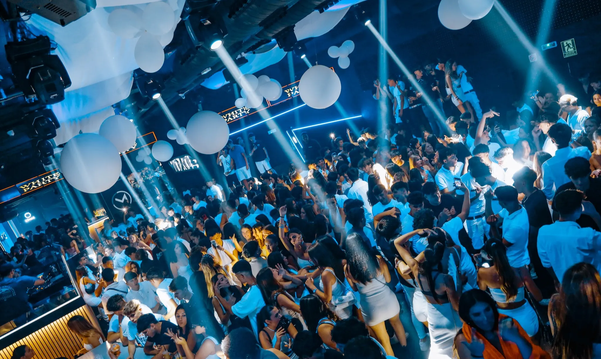 Discoteca llena de gente vestida de blanco bajo luces azules y globos gigantes en el techo durante una fiesta en Miracle Mataró.