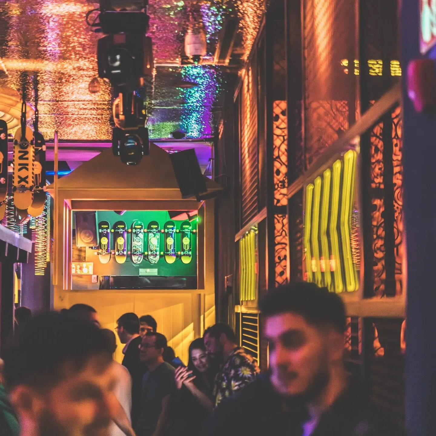 Imagen de HYP3 conambiente nocturno y animado en un club, con luces de colores que crean un efecto brillante en el techo. Varias personas socializan y se ven detalles de decoración, como monopatines en la pared.