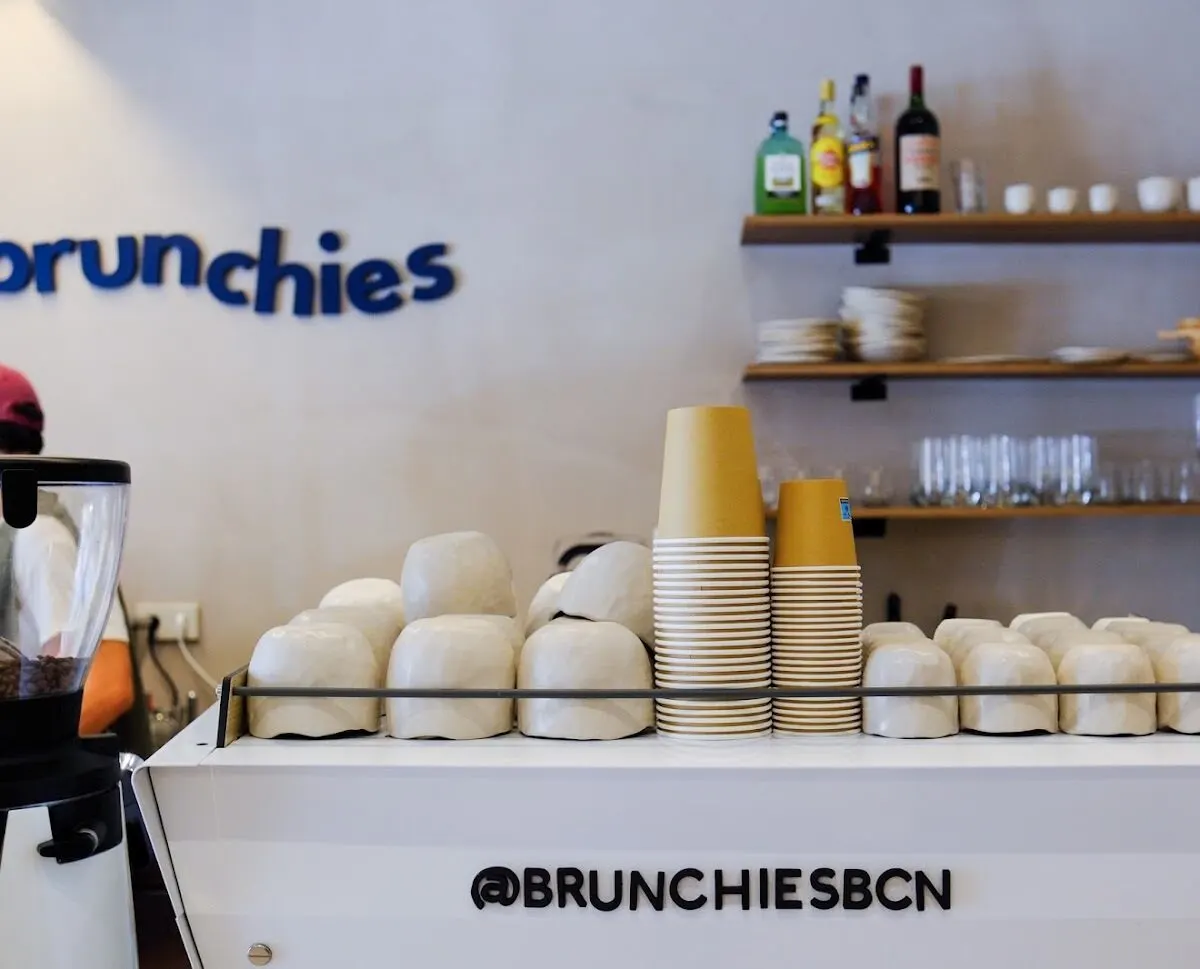 Mostrador de Brunchies con máquina de café blanca, vasos de cartón apilados, tazas blancas y estanterías con botellas y cristalería al fondo.