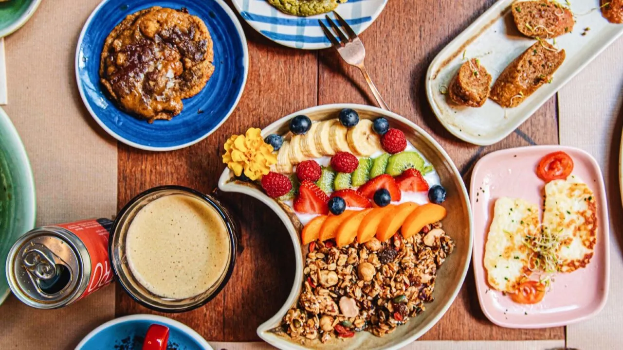 Desayuno en Faire Brunch & Drinks con bol de yogur y frutas frescas, galleta, pan, queso a la plancha, café y refresco sobre mesa de madera.