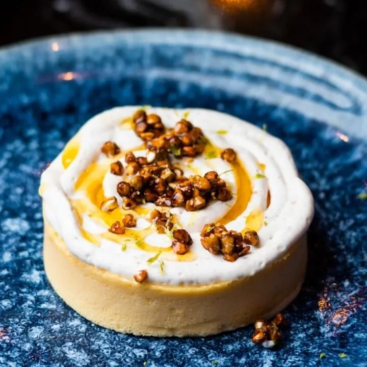 Tarta sobre plato cerámico azul, con base clara, crema batida, salsa de caramelo y frutos secos crujientes como decoración.