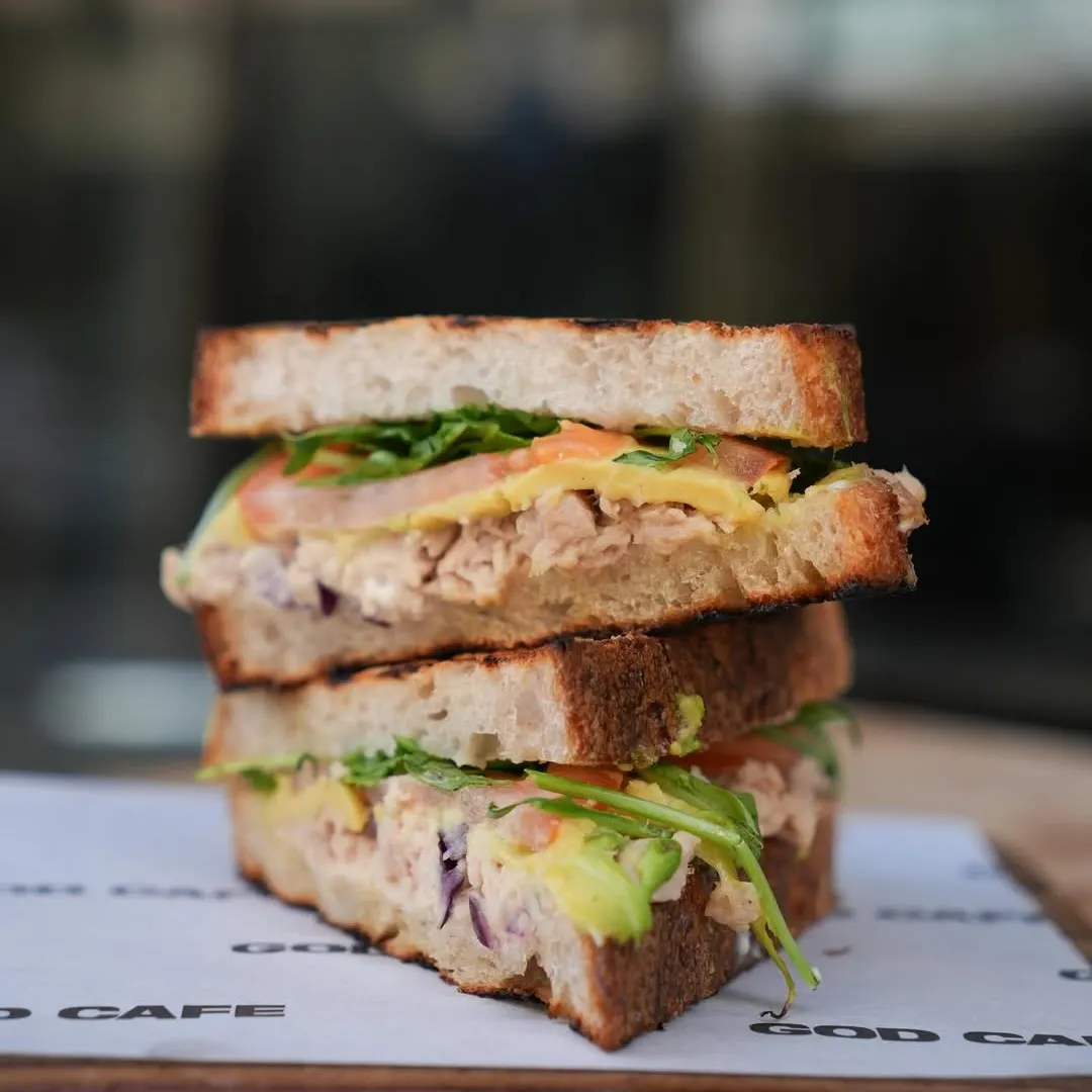 Sándwich de God Café con pan tostado relleno de aguacate, tomate, rúcula, queso y pollo desmenuzado servido sobre papel con el nombre del local