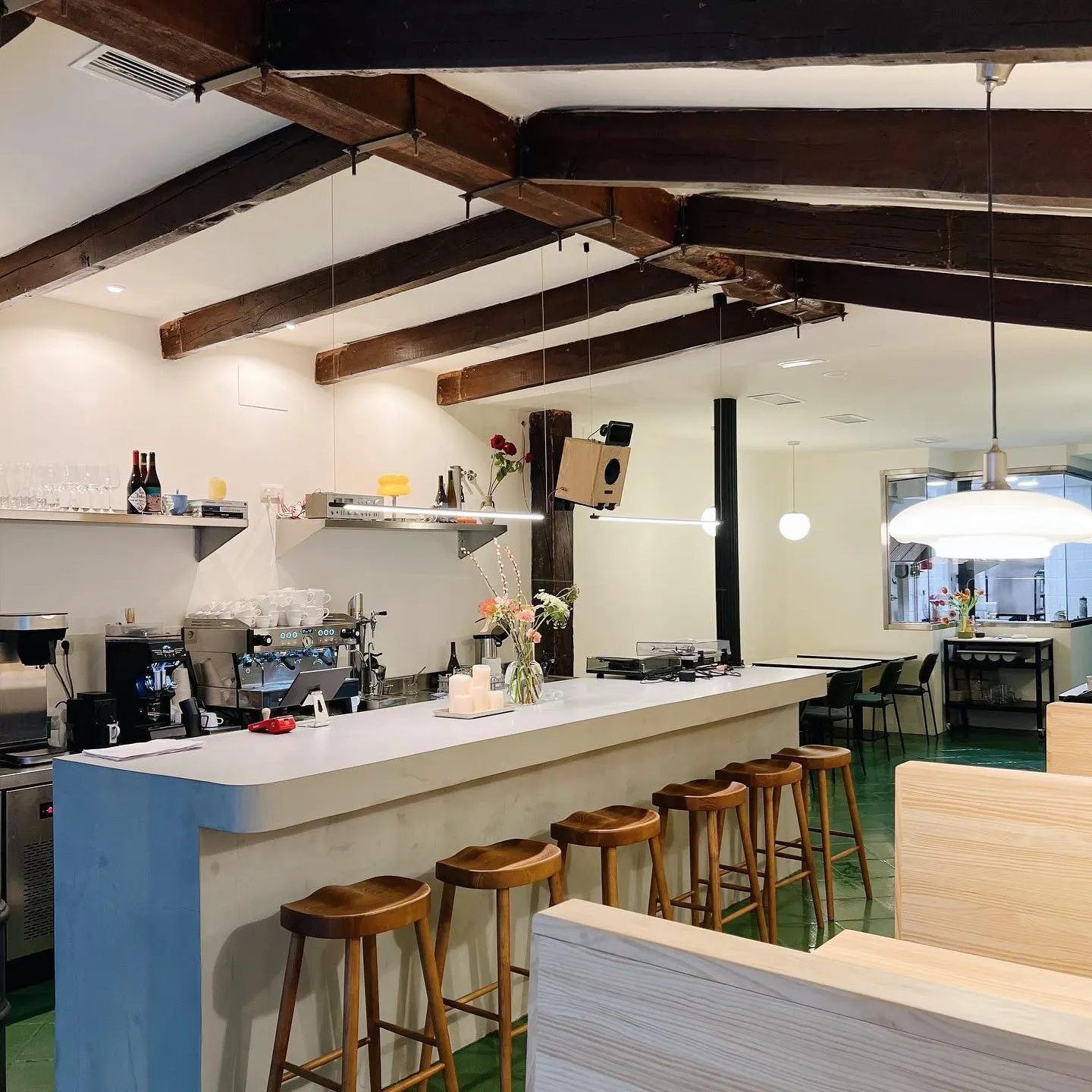 Interior de cafetería moderna con barra larga, taburetes de madera, flores, velas, estanterías con botellas y techo con vigas vistas.