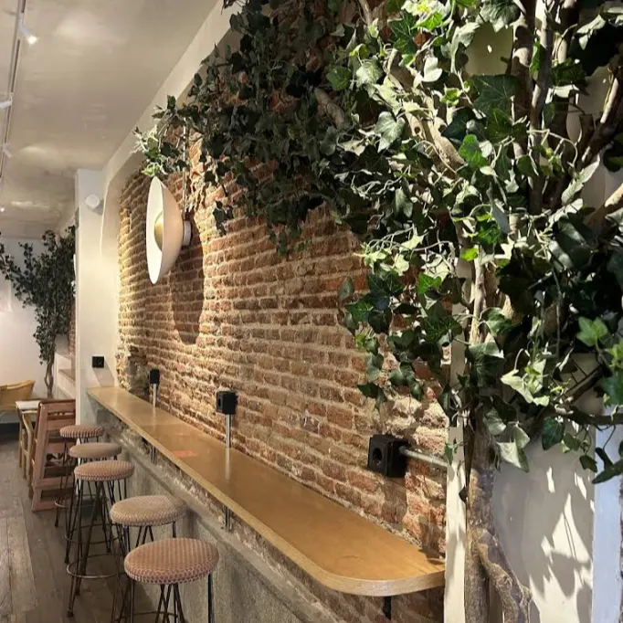 En Maison Kayser Quevedo hay una barra de madera frente a una pared de ladrillo con plantas decorativas y taburetes tapizados.