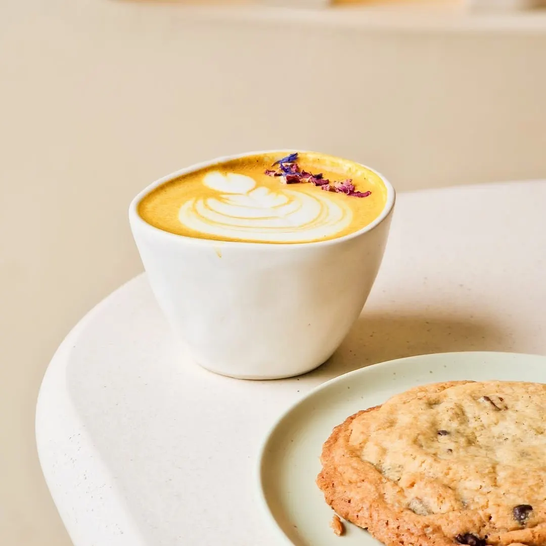Latte dorado con arte en la espuma y pétalos secos, acompañado de una cookie con chips de chocolate sobre un plato verde pastel en una mesa de tono claro.