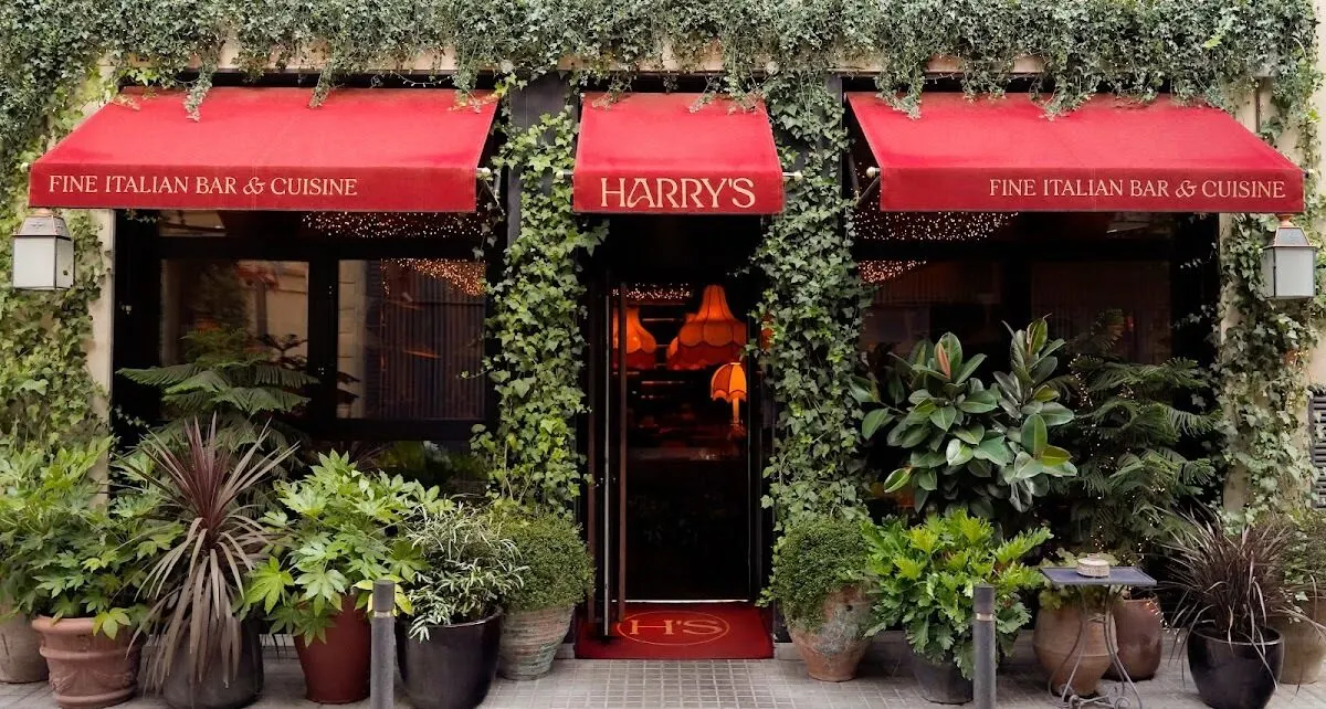 Entrada del restaurante “Harry’s” con fachada cubierta de hiedra, toldos rojos y plantas en macetas. Estilo elegante con aire italiano.