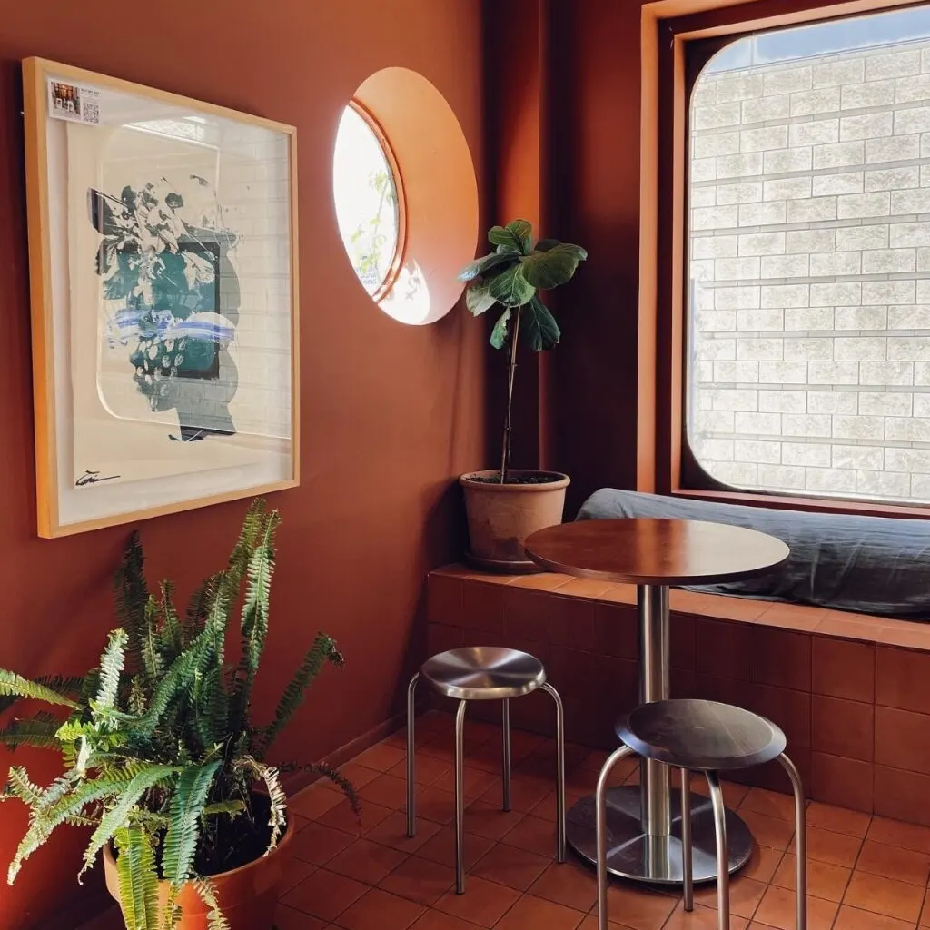 Esquina cálida con paredes marrón, suelo de baldosas, ventana redonda y rectangular, plantas verdes y mesa metálica con dos taburetes.