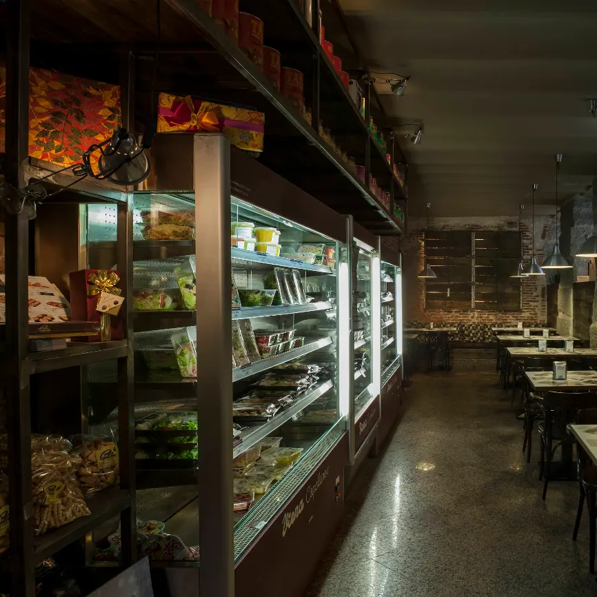 Zona de mesas en Viena Capellanes Ruiz de Alarcón, con neveras de platos preparados, estanterías con productos gourmet y decoración rústica e industrial.