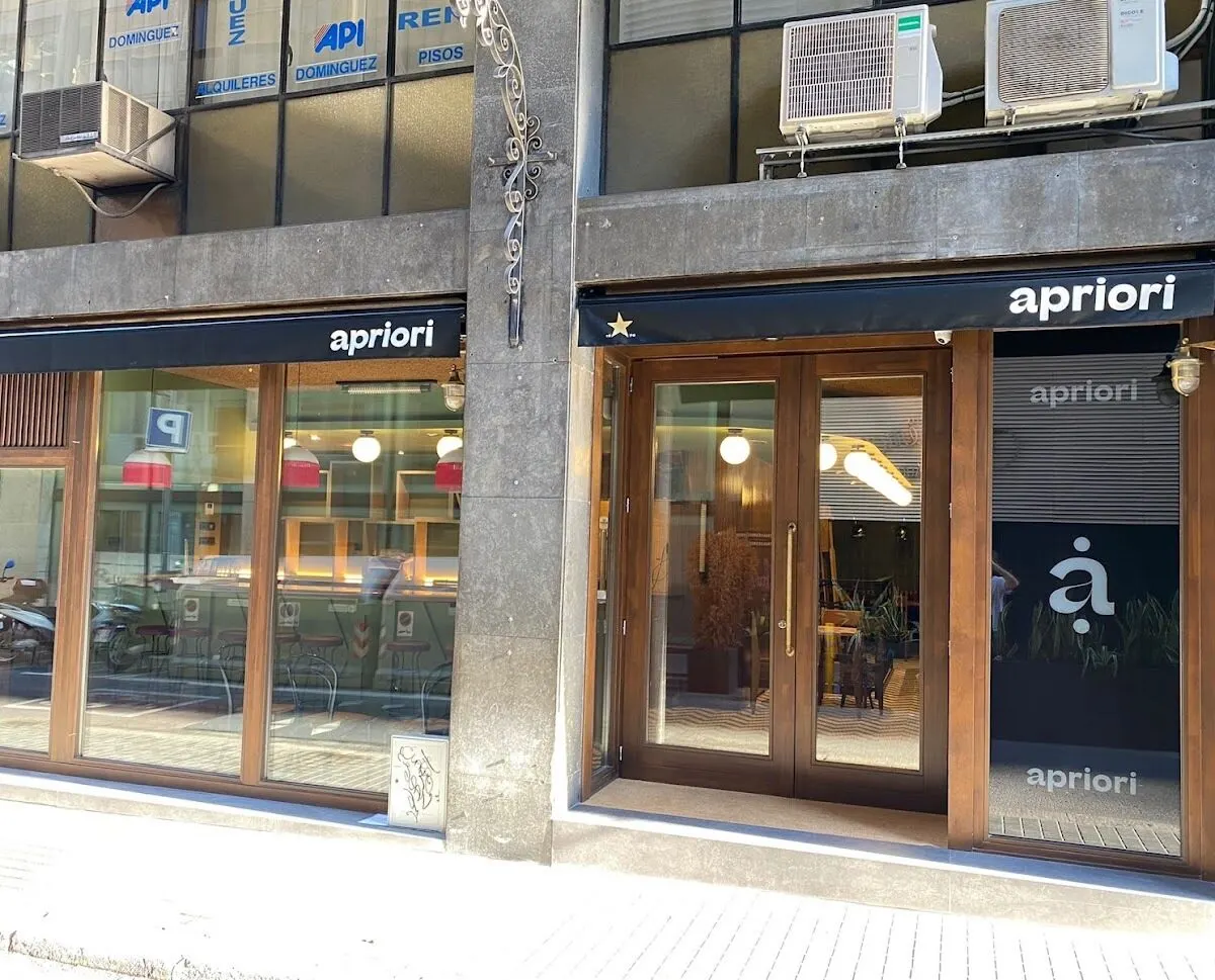 Fachada de edificio con ventanas grandes y dos entradas bajo el cartel “apriori”. Interior visible con mesas, sillas y luces. Aires acondicionados arriba.