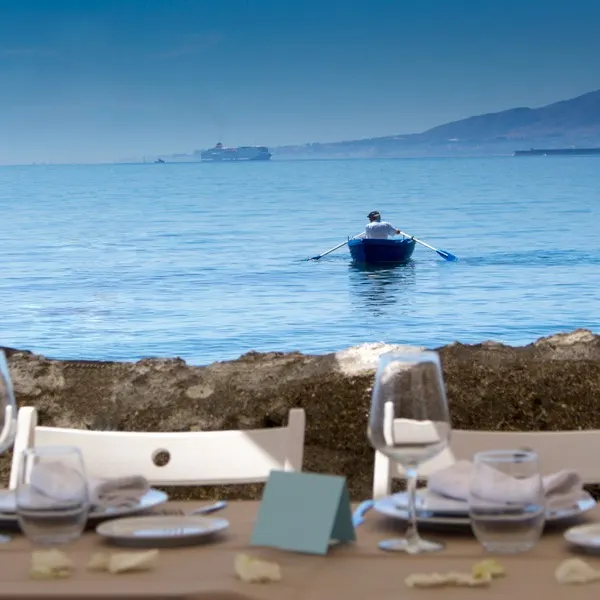 La imagen muestra una mesa de comedor en El Balneario, preparada para una comida con vasos transparentes, platos blancos y servilletas dobladas, con vista a un cuerpo de agua sereno. A lo lejos, hay una persona remando en un pequeño bote azul.