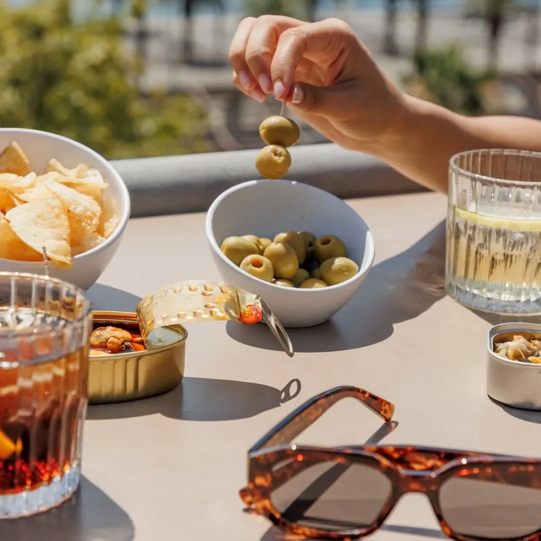 Aperitivo en la terraza de La Duquesa de Cardona con vistas al puerto: vermut, aceitunas, patatas y conservas al sol, junto a unas gafas de sol sobre la mesa.