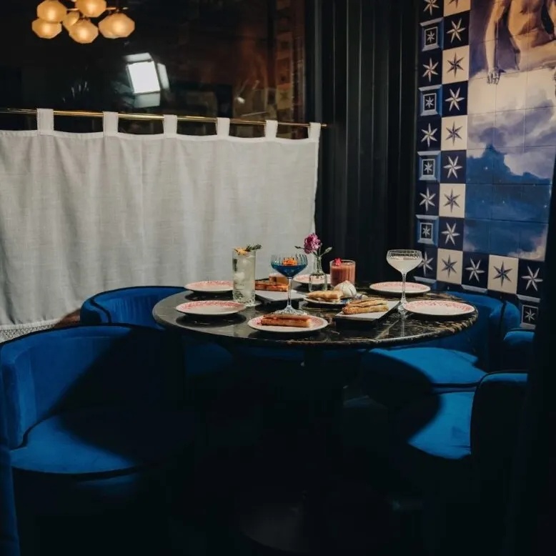 Mesa redonda con cuatro sillas azules, platos servidos, copas y lámpara moderna; fondo con mural artístico y cortina blanca.