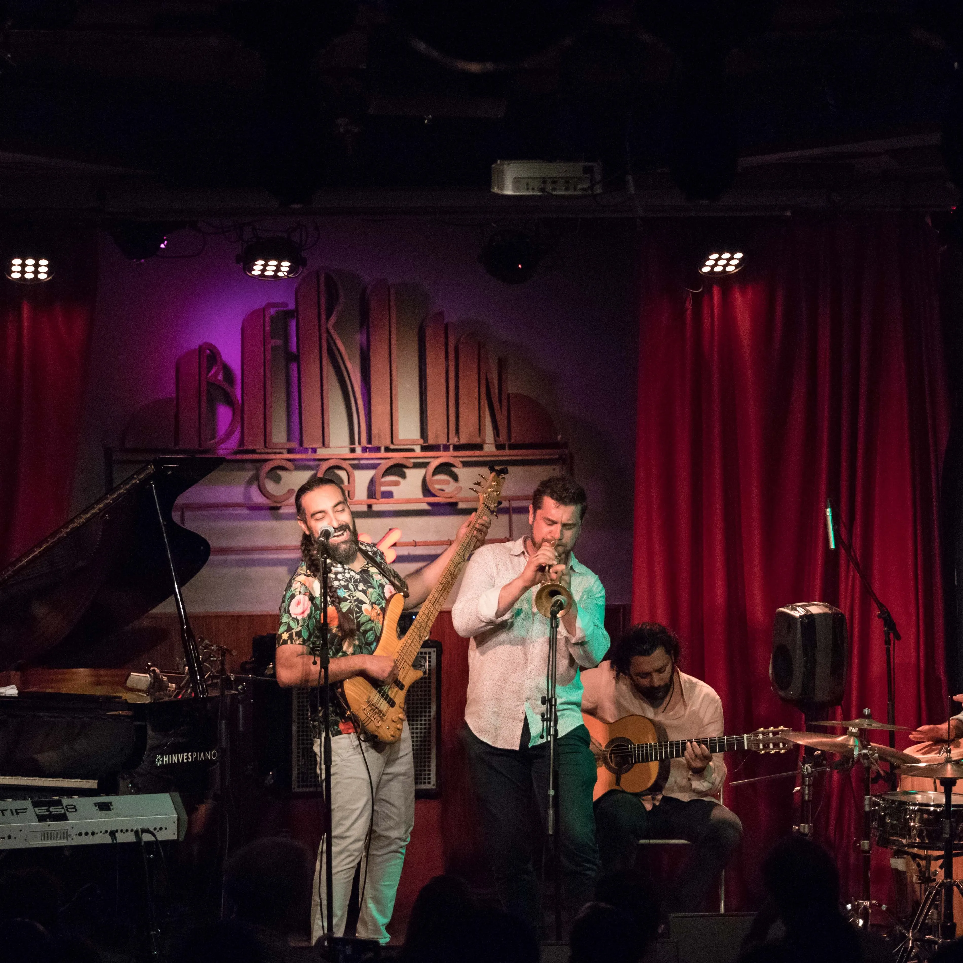 Banda en vivo actuando en el escenario del Café Berlín, rodeados de cortinas rojas y un ambiente íntimo con luces cálidas.