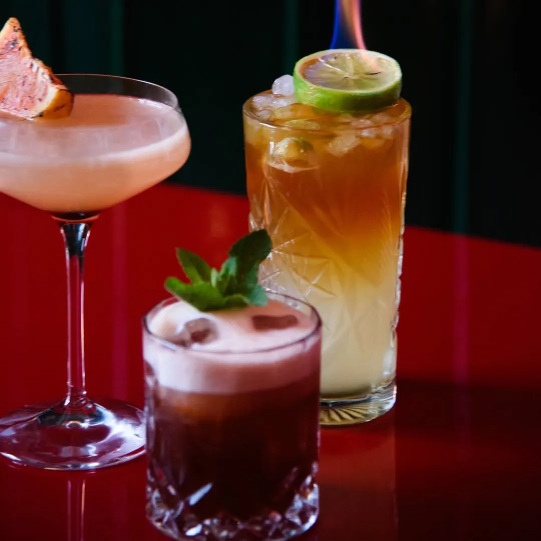 Tres cócteles sobre mesa roja: uno en copa de cóctel con piel de cítrico, otro oscuro con espuma y uno tropical con lima flameada.