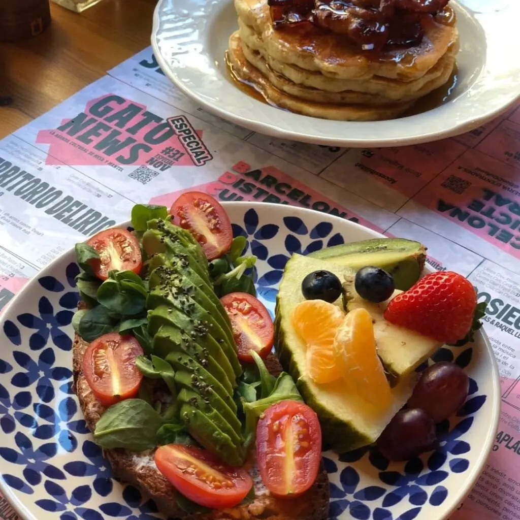 Tostada con aguacate, frutas y queso junto a pancakes con mantequilla y sirope sobre mantel tipo periódico.