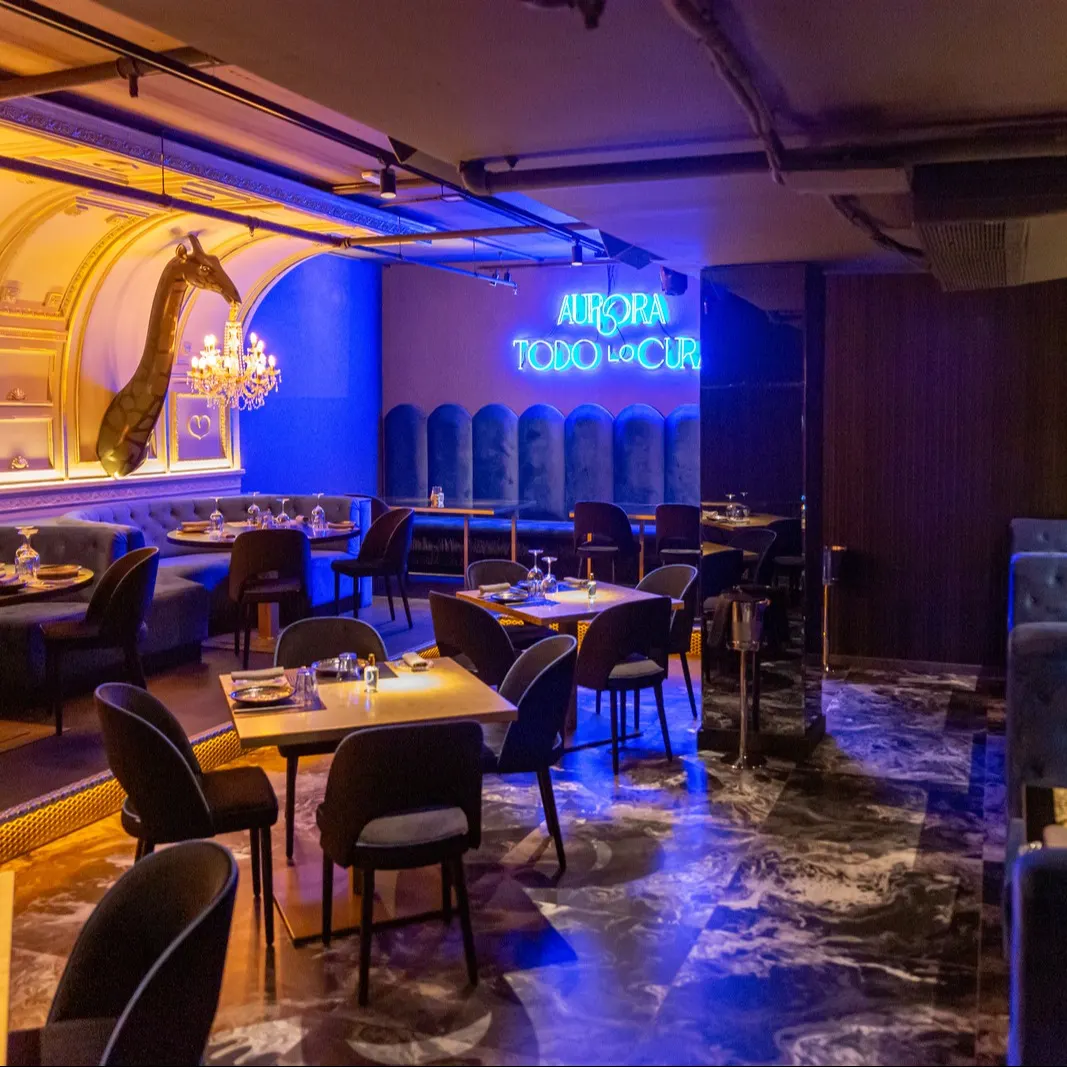 Un elegante comedor en Aurora con paredes azules, mesas de madera y detalles únicos como jirafas decorativas y un neón que dice "Todo lo cura".