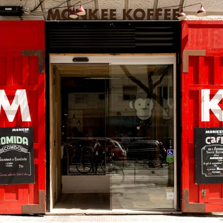 Entrada de cafetería con puertas rojas marcadas con “M” y “K”. Pizarras anuncian combos y café arábica. Interior visible tras puerta de cristal.