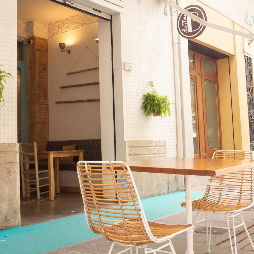 La Más Bonita luce una fachada mediterránea con detalles en azul pastel, sillas de mimbre y una atmósfera acogedora perfecta para cualquier hora del día.