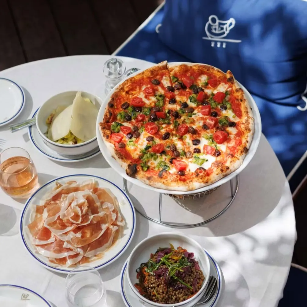 En Cipriani, una pizza fresca con tomates, aceitunas y pimientos se acompaña de jamón, ensalada y una copa de vino rosado para un almuerzo perfecto.