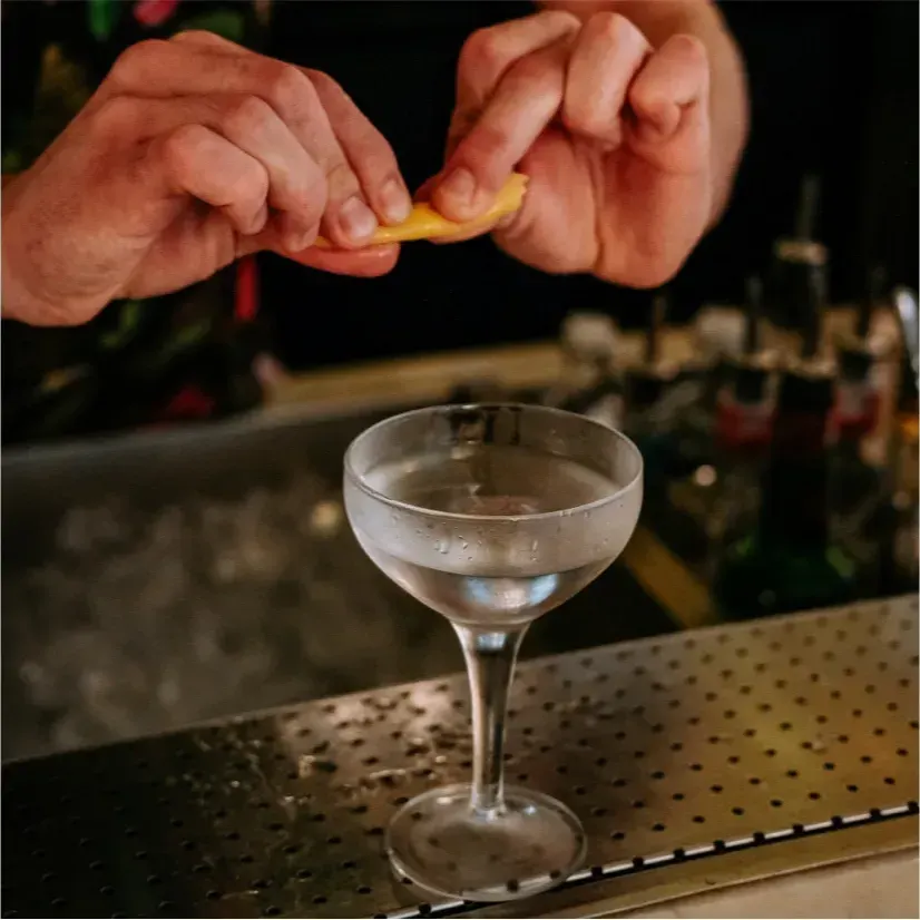 Manos exprimen piel de cítrico sobre cóctel transparente en copa coupe; fondo de bar con botellas y utensilios visibles.
