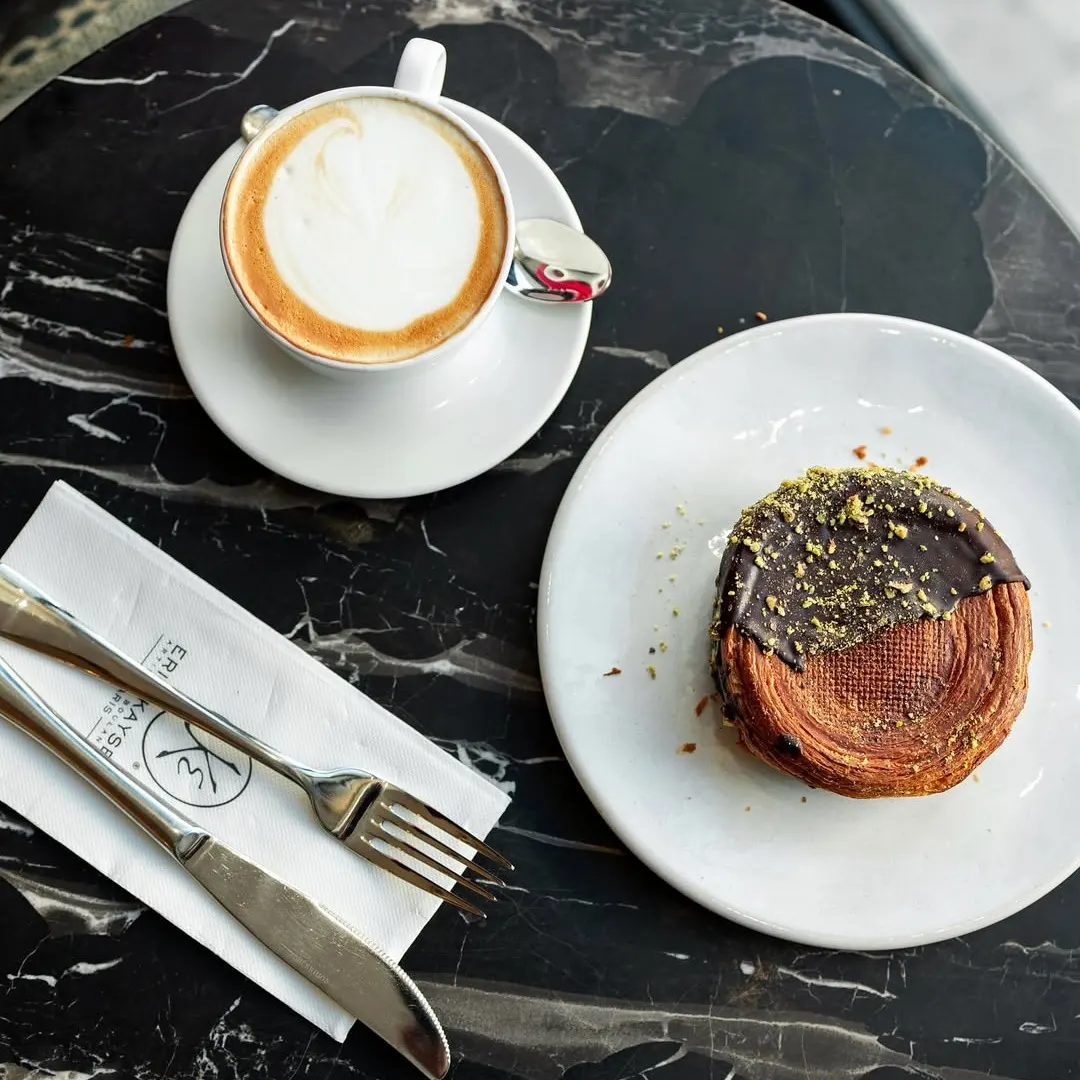 Café con espuma servido junto a un pastel hojaldrado con cobertura de chocolate y pistacho en una mesa de mármol negro en Maison Kayser.
