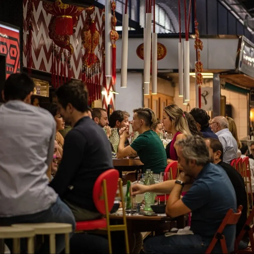 Zona de comedor interior con varias personas comiendo; decoración con faroles rojos y adornos colgantes en ambiente de mercado o restaurante.