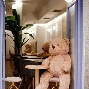 Cafetería interior con osos de peluche en mesas, luz cálida, plantas decorativas y lámparas modernas que crean un ambiente acogedor.