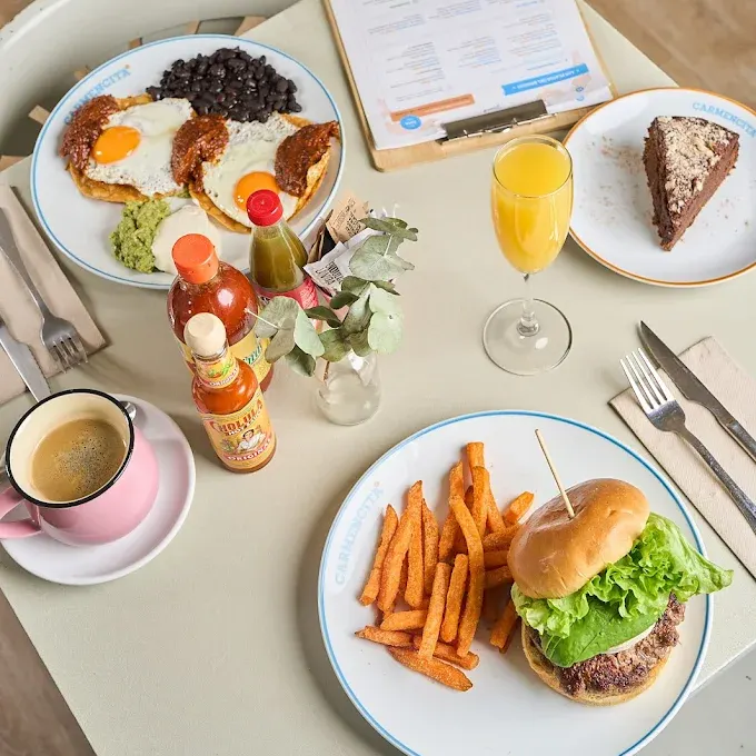 Mesa con hamburguesa y boniato, huevos con guacamole y salsa, pastel de chocolate, zumo de naranja, café rosa y botellas de salsa picante.