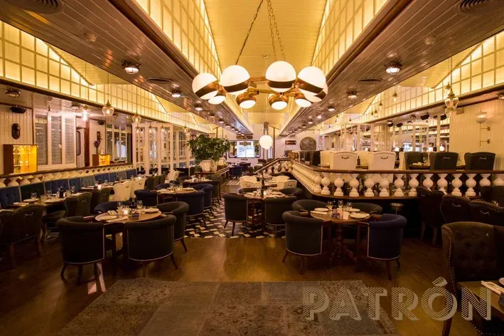 La foto muestra el interior de Patron. El techo está adornado con múltiples lámparas que proporcionan una luz cálida al espacio. A la derecha, hay una zona de bar con taburetes y estantes llenos de varias botellas.