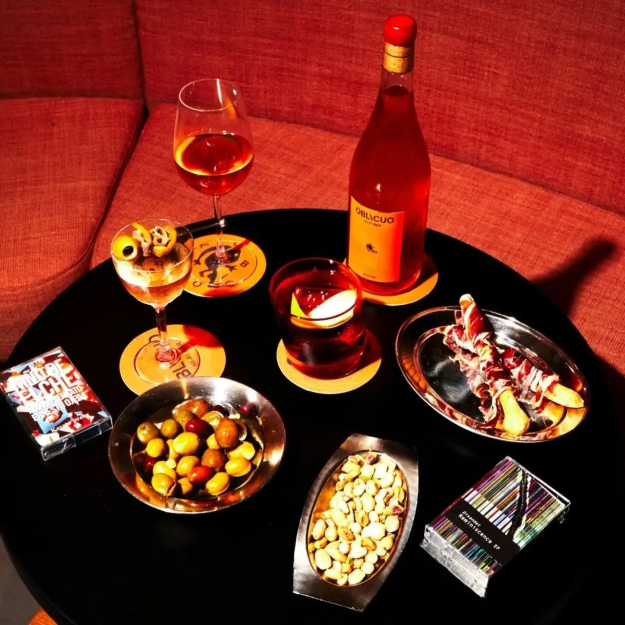 Mesa negra con vino Oblicuo, copas, aceitunas, frutos secos, embutidos, bebida oscura y dos barajas de cartas.
