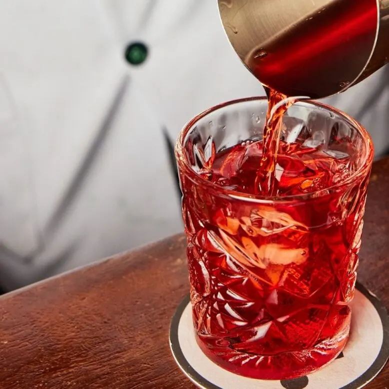 Persona con prenda blanca y botones verdes sirve líquido rojo en vaso decorado, sobre posavasos en mesa de madera.