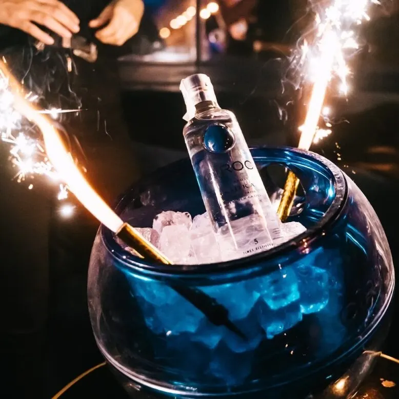 Botella de vodka Ciroc en cubo azul con hielo y bengalas encendidas; fondo con personas desenfocadas.