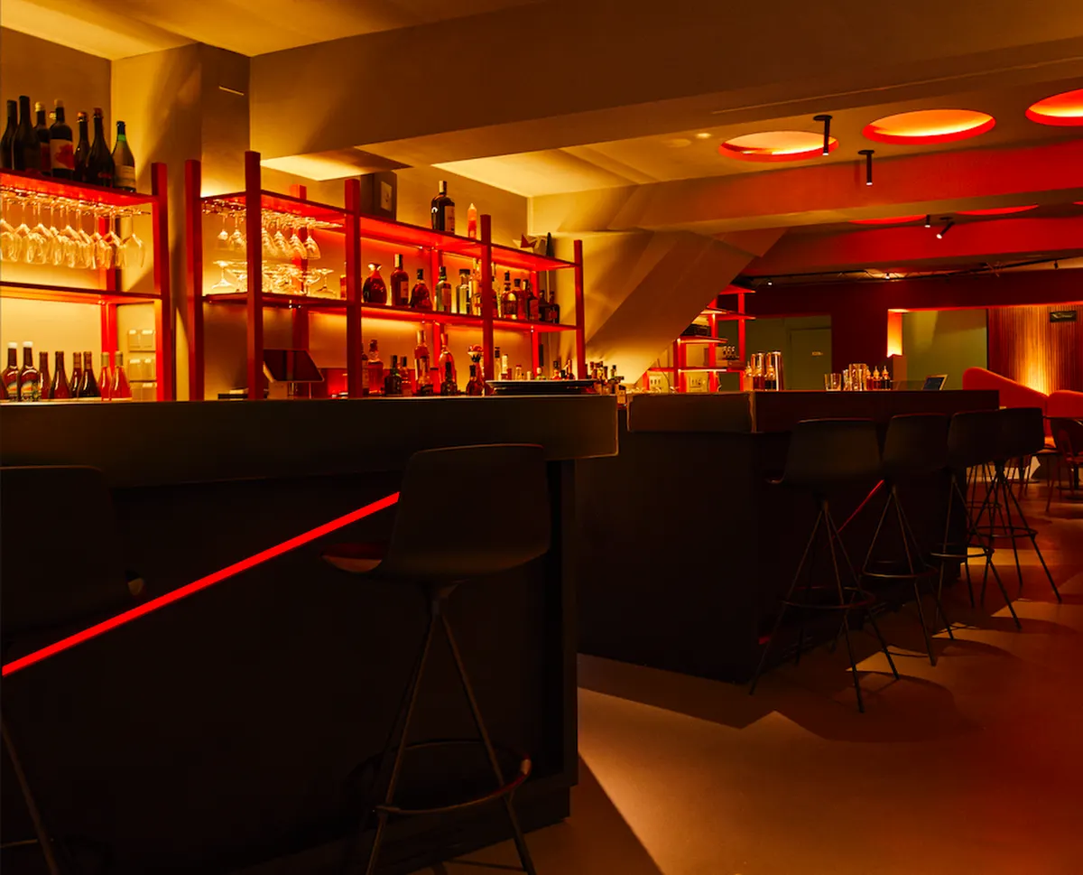 Bar moderno con iluminación cálida, estanterías con botellas y copas, y sillas altas en la barra. Ambiente acogedor y diseño contemporáneo.