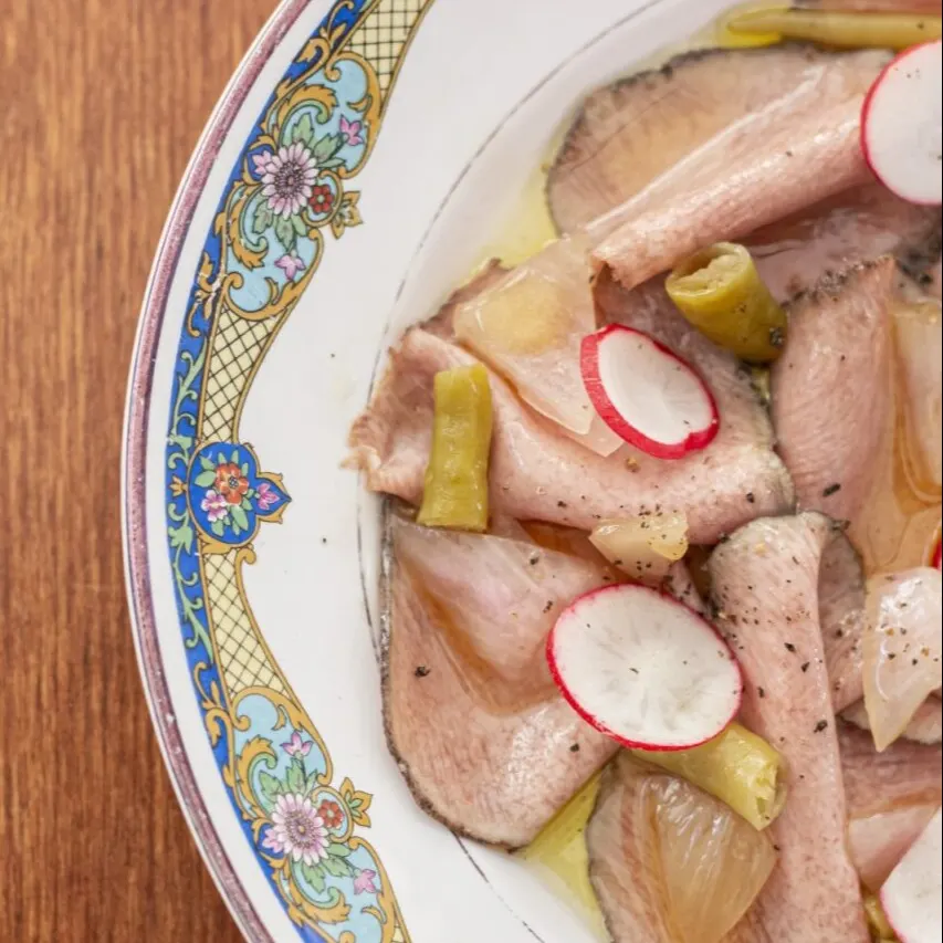 Plato decorativo con patrón floral y geométrico, contiene pescado en láminas con rábano, pimientos verdes y cebolla sobre fondo de madera.