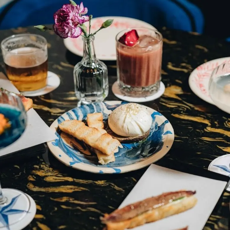 Mesa con pasteles, postres con crema y bebidas, una con flor rosa. Jarrón pequeño con flor morada sobre superficie de mármol oscuro.
