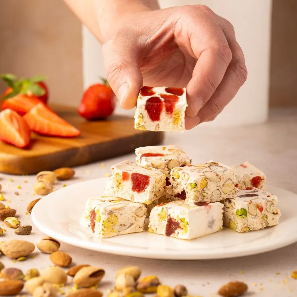 Cuadrados de turrón blando con pistachos y trozos de fresa confitada sobre un plato blanco, rodeados de frutos secos, con una mano destacando una pieza en primer plano.