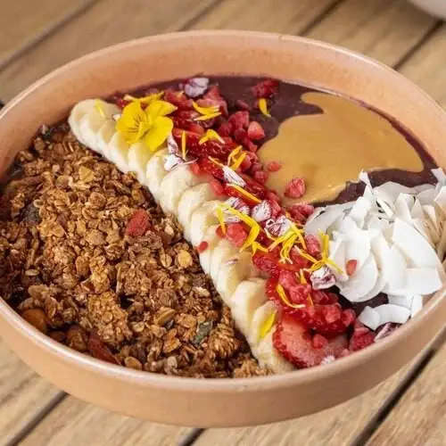 Bol de açaí con granola, plátano, fresas, coco, mantequilla de cacahuete, flores comestibles y bayas, todo bien ordenado.