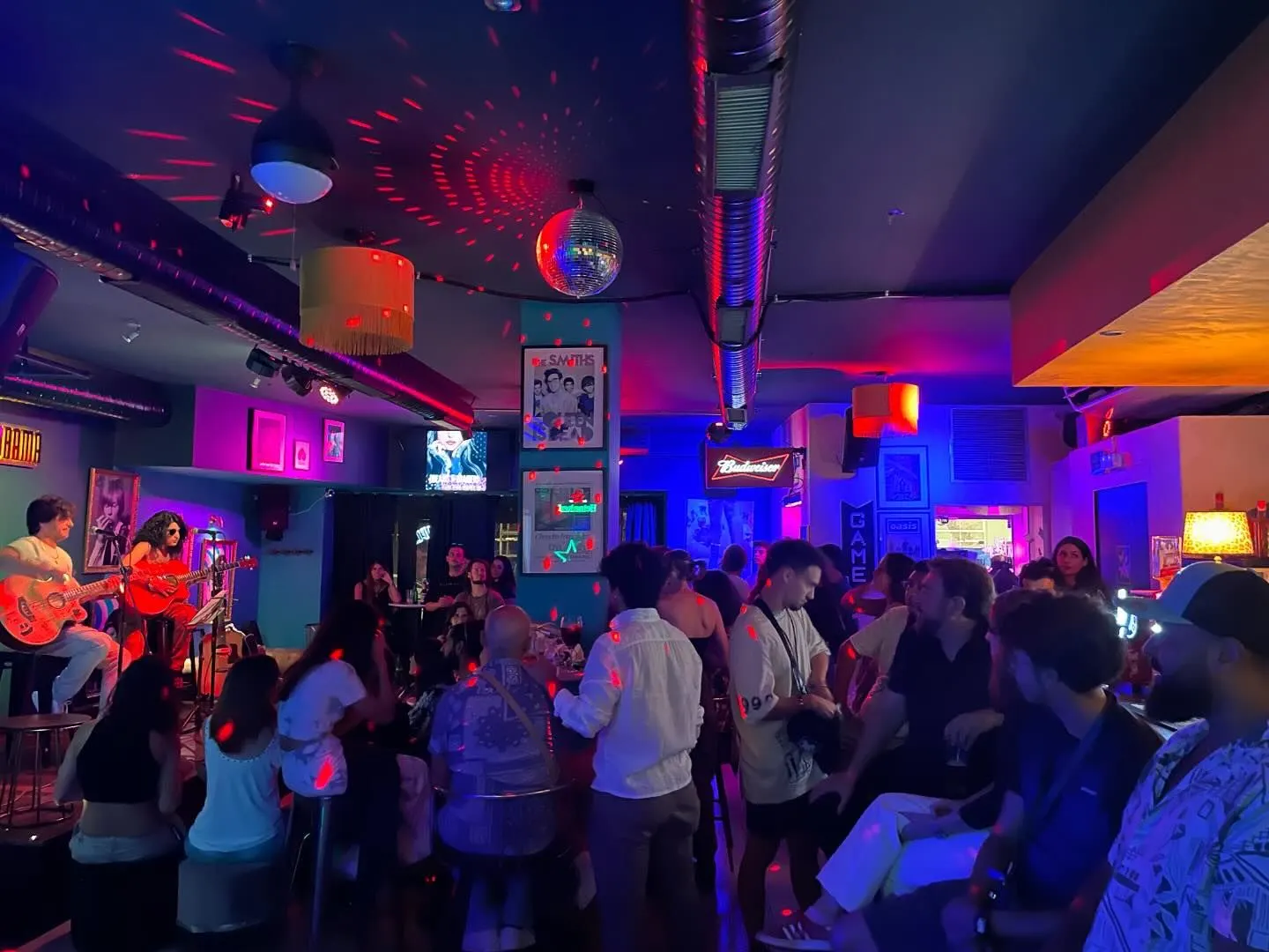 " La imagen muestra una presentación musical en Lady Drama, un bar con iluminación de tonos morados y azules, decorado con un ambiente retro y posters icónicos. Los asistentes disfrutan del show en una atmósfera vibrante y acogedora."