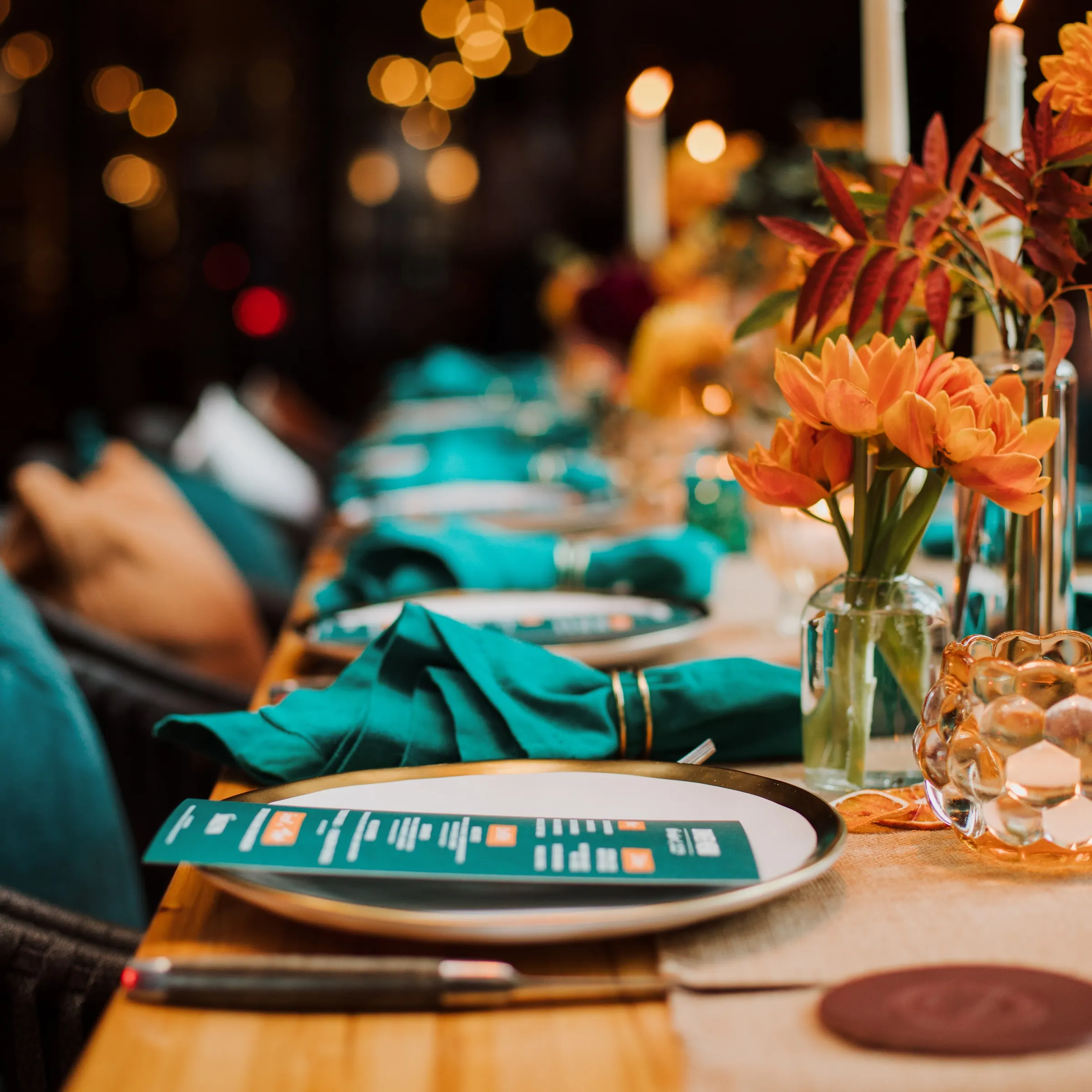 Mesa festiva elegante dispuesta con servilletas azules, platos decorativos y arreglos florales naranjas, bajo la cálida luz de las velas.