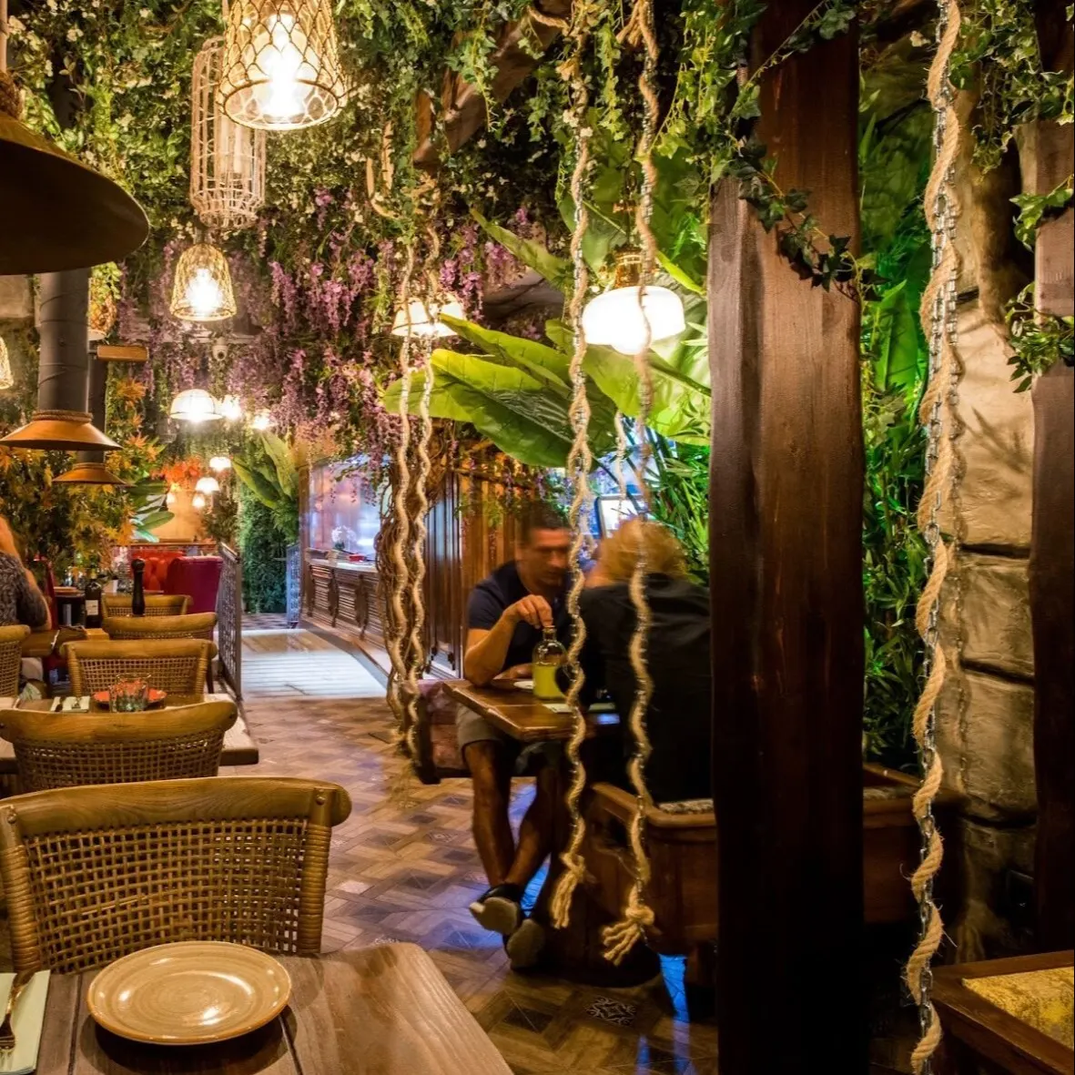 Restaurante interior con techo cubierto de plantas colgantes, mesas de madera, luz cálida y ambiente selvático acogedor.