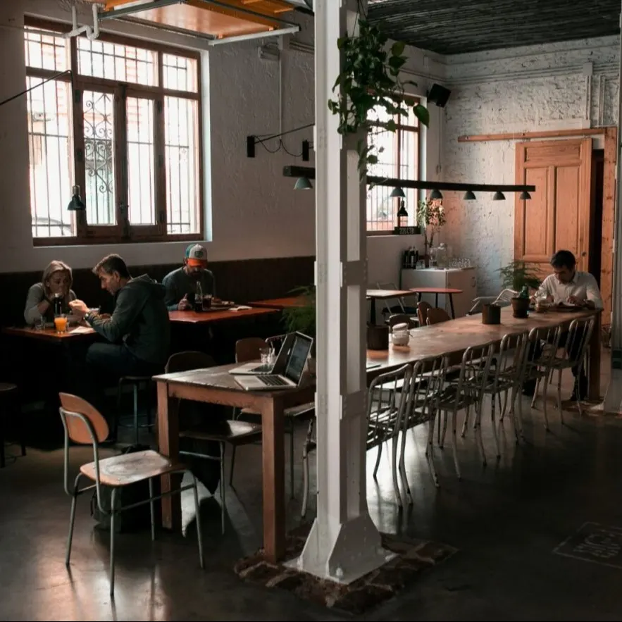 Interior de cafetería con mesas ocupadas, ventanas con rejas metálicas, plantas colgantes y luz cálida que crea ambiente acogedor.