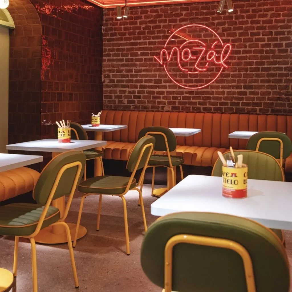 Interior moderno con mesas blancas, sillas verdes con marco amarillo y bancos naranjas. Latas con cubiertos y neón “mazál” sobre pared de ladrillo.