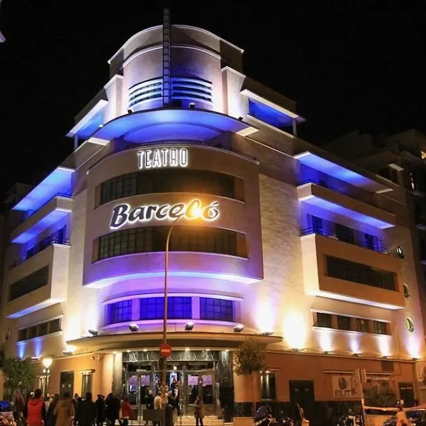Edificio iluminado de noche con estilo art déco. Letrero “TEATRO Barceló” en fachada. Luces azules y púrpuras. Personas reunidas en la entrada.