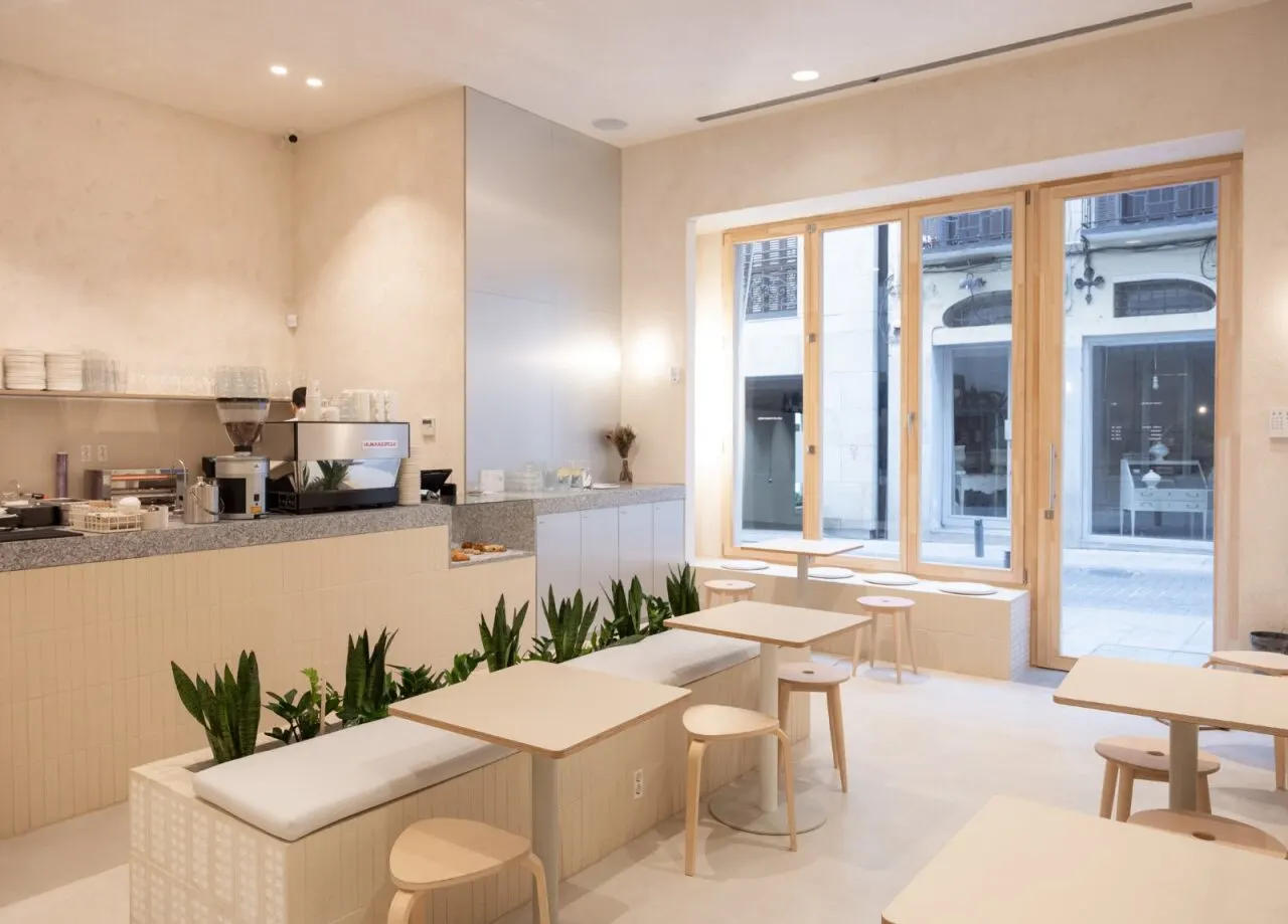 Interior luminoso de Casa Neutrale con mesas claras, bancos con plantas y una barra de café con máquina La Marzocco junto a una gran ventana.