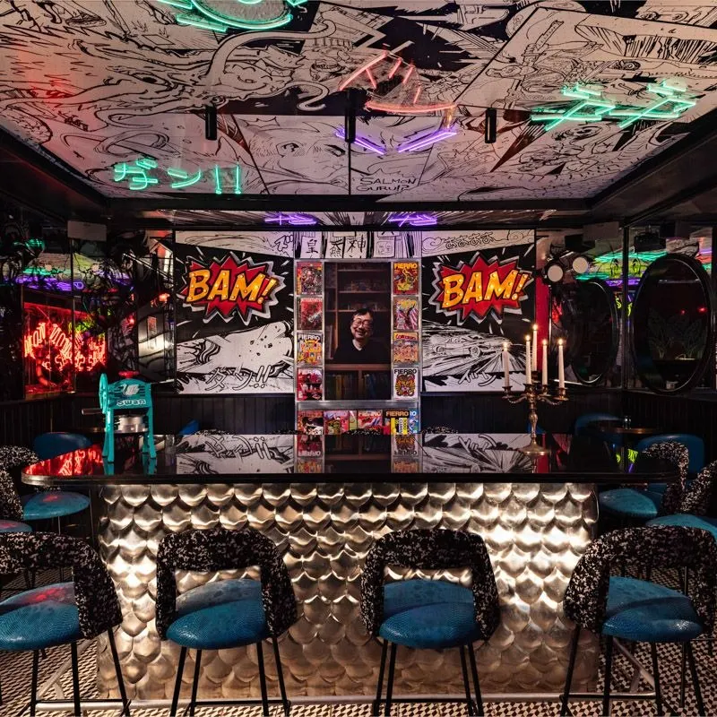 Barra metálica rodeada de sillas azules con respaldo estampado, decorada con murales de cómic y luces de neón en el techo.