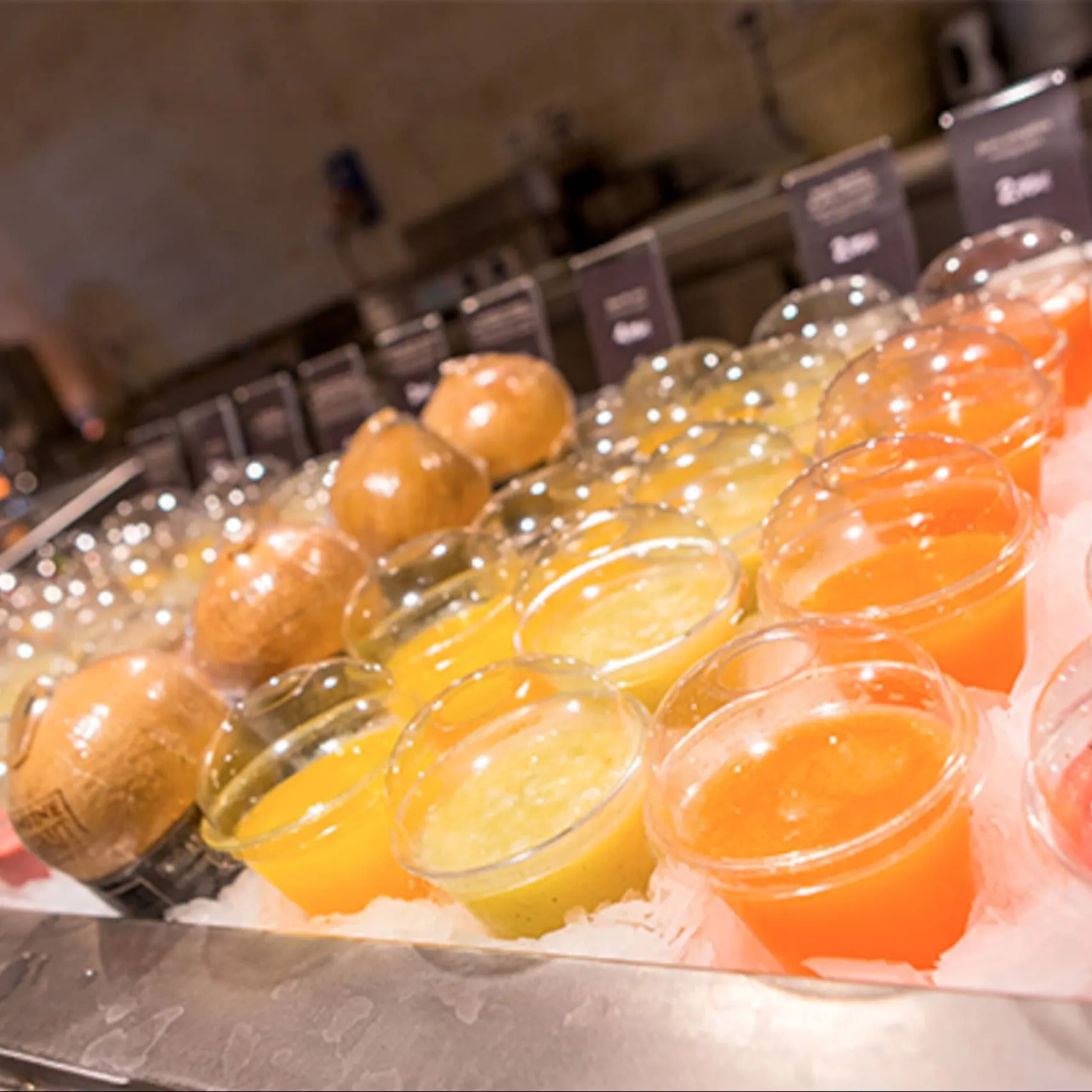 Vasos de zumo natural de distintos sabores sobre una bandeja de hielo picado, con carteles de precios al fondo y cocos enteros entre ellos.