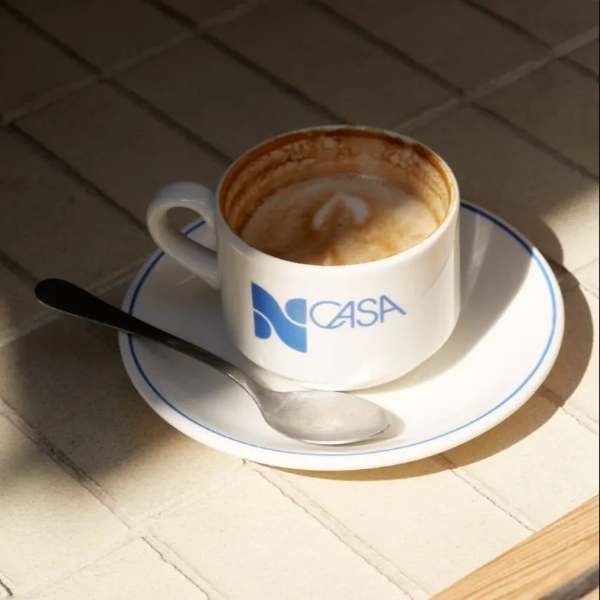Taza blanca con logo azul “CASA” sobre platillo blanco con borde azul y cuchara metálica; fondo de baldosas con luz solar.