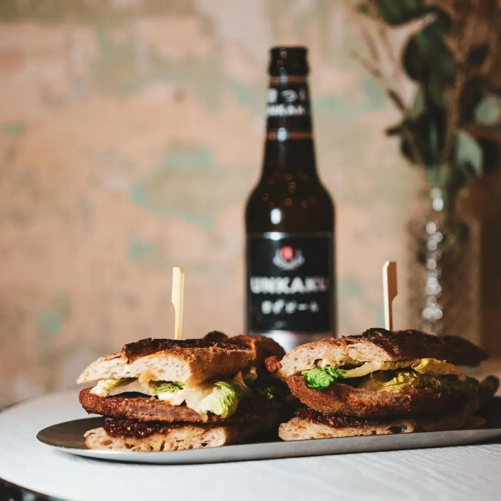 Dos sándwiches con palillos en plato, botella de cerveza Unkari al fondo, pared desenfocada y plantas decorativas en jarrón.