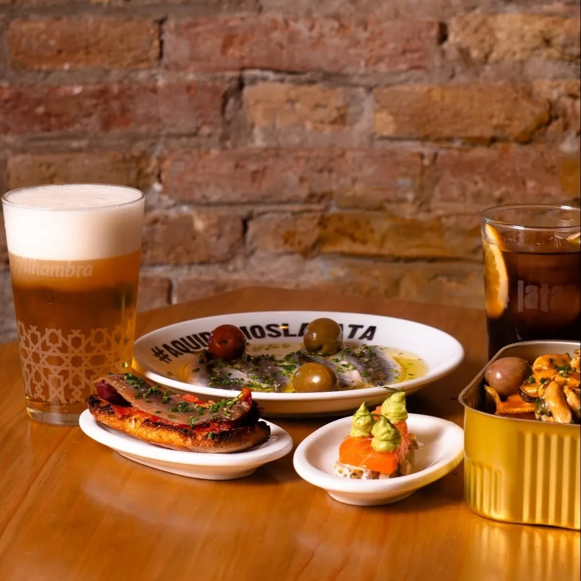 Mesa con cerveza, refresco, tapas de marisco, salmón y carne; fondo de ladrillo y plato central con texto “#AQUIHAYOSLATA”.
